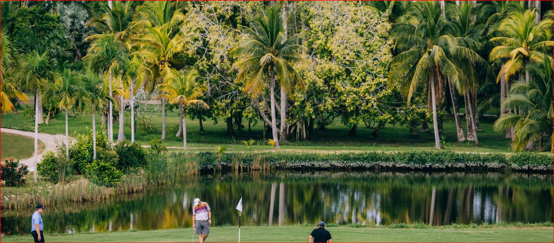 Golfers play on a green at Balam Golf Course, surrounded by palm trees, tropical landscaping, and a calm water feature near the Placencia Lagoon.