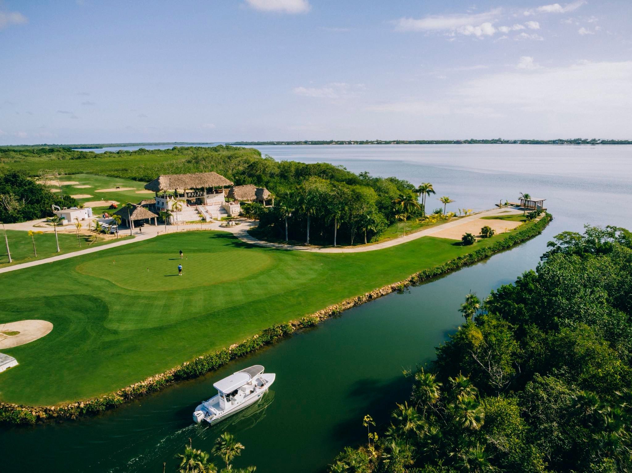 Aerial view of Balam Golf Course beside the Placencia Lagoon, with a boat approaching the docking area and the golf course, clubhouse, and waterfront landscape visible from above.