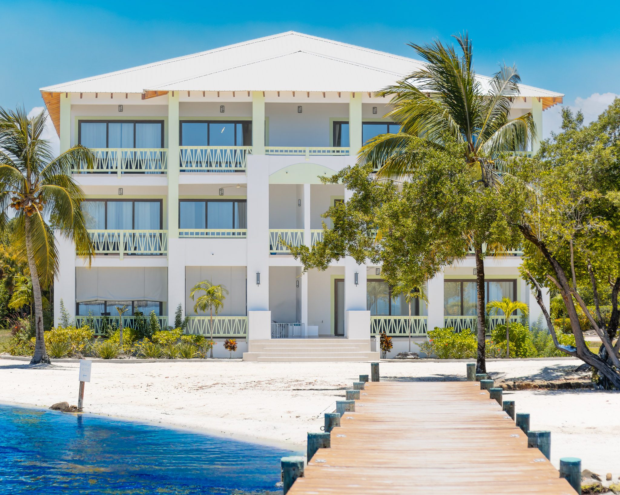 Marazul Belize beachfront property viewed from the private pier, with a white modern coastal building, palm trees, turquoise water, and a clear blue sky in the Surf Side area of the Placencia Peninsula.